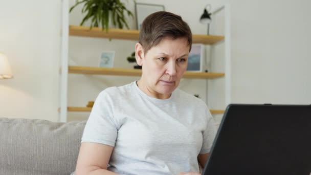 Χαμογελαστή ηλικιωμένη ώριμη γυναίκα ξεκουράζεται στον καναπέ, χρησιμοποιώντας το φορητό υπολογιστή μόνη στο σπίτι. Καλή παλαιότερη συνταξιούχος web surfing πληροφορίες. Σπουδές ή online αγορές — Αρχείο Βίντεο