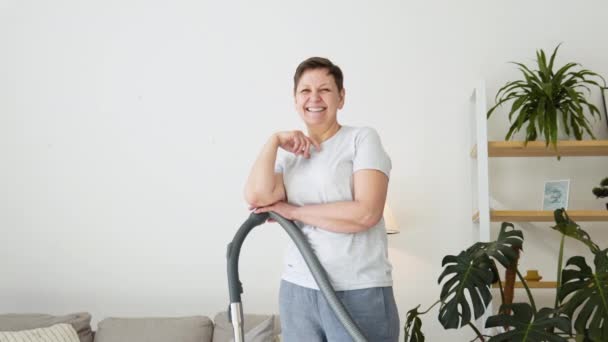 Portrait of a senior woman with a vacuum cleaner while cleaning. Housekeeping routine. Domestic hoover appliance cleaner — Vídeos de Stock
