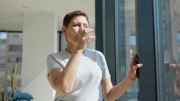 Senior vrouw drinkt water staande in de buurt van het raam in een lichte kamer — Stockvideo