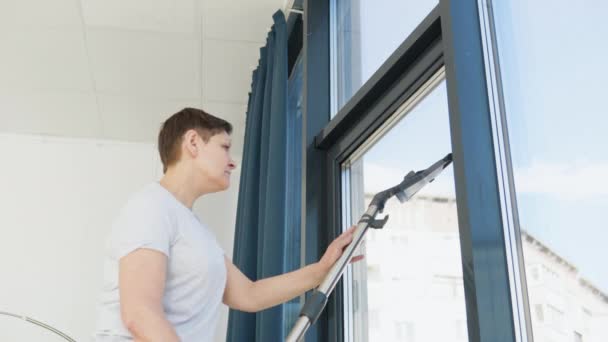 Senior woman cleaning window at home with a vacuum cleaner. Professional cleaning woman during work — Vídeos de Stock