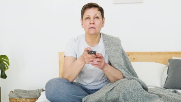 Senior lady watching tv in living room sitting on sofa holding remote control changing tv channels. Senior woman using TV remote control — Vídeo de stock