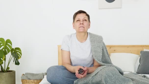 Senior lady watching tv in living room sitting on sofa holding remote control changing tv channels. Senior woman using TV remote control — Vídeo de stock
