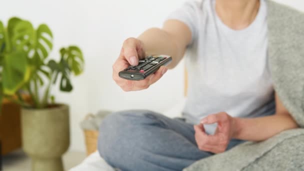 Senior lady watching tv in living room sitting on sofa holding remote control changing tv channels — Vídeos de Stock