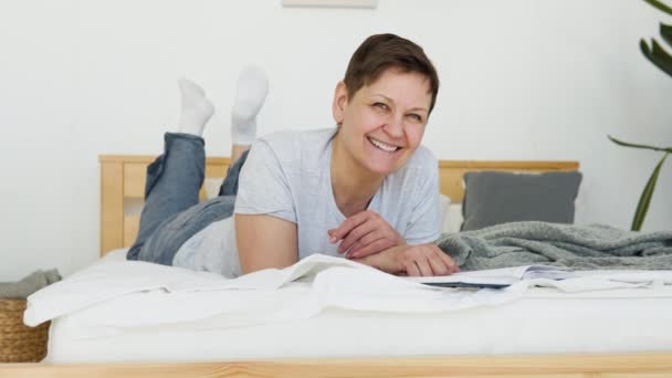 Relaxed senior woman lying on bed and reading book indoors at home. Leisure activity of mature female at comfort modern flat. Casual elderly life and wrinkled skin closeup — стоковое видео