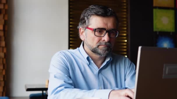 Vista lateral del hombre mayor que se sienta en la cafetería durante el día y el uso de la computadora portátil para el trabajo remoto — Vídeo de stock