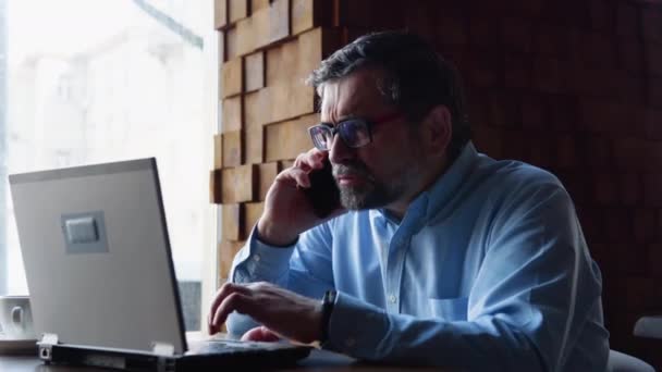 Großaufnahme eines leitenden Geschäftsmannes, der mit dem Handy telefoniert und im Café auf den Laptop schaut — Stockvideo