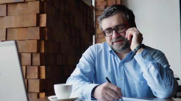 Крупный план старшего бизнесмена, разговаривающего по мобильному телефону, смотрящего на ноутбук в кафе — стоковое видео