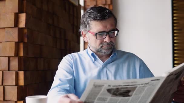 Senior man reads a newspaper sitting in a cozy cafe — Vídeo de stock