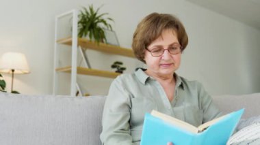 Gülümseyen son sınıf öğrencisi kadın kitap okuyarak rahatlıyor evde kanepede oturuyor.