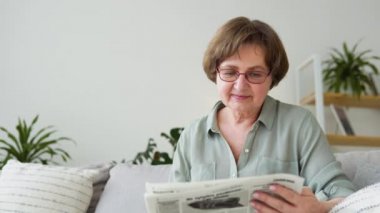 Gözlüklü yaşlı kadın evde gazete okuyor.
