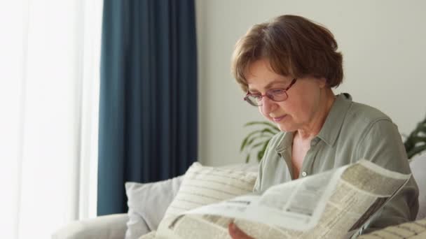Senior woman with glasses reads newspaper at home — Video