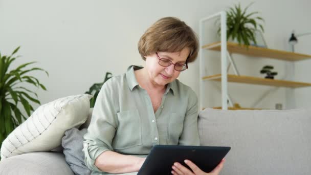 Улыбающаяся пожилая женщина отдыхает на диване, используя цифровые планшеты дома — стоковое видео