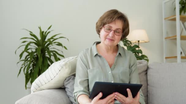 Bella signora anziana guardando in macchina fotografica seduta in soggiorno con tablet — Video Stock