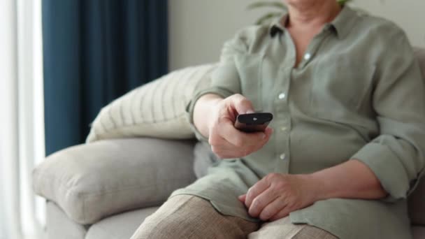 Aged woman watching tv in living room sitting on sofa holding remote control changing tv channels — Vídeo de stock