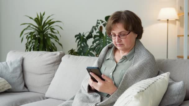 Старшая бабушка пользоваться мобильным приложением сидеть на диване — стоковое видео