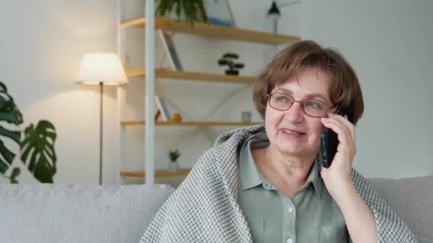 Šťastný senior žena relaxovat na útulné pohovce, mluvit na smartphone s přáteli — Stock video