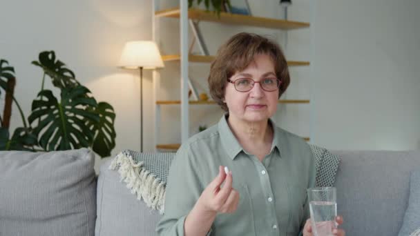 Beautiful senior woman with a pill and a glass of water in her hands sits on a sofa at home — Vídeo de stock