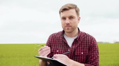 Portrait shot of attractive farmer standing in green field with green wheat and notebook in the hands. Farmer with smile outdoors in summer