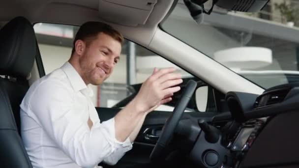 Portrait of a happy young man driving a new luxury electric car in a car dealership. The man shows emotions of happiness while driving in her new car. The buyer selects a car — Stock Video