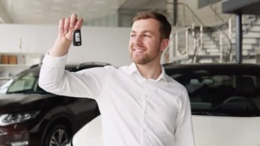 Portrait of happy adult successful man posing in auto showroom buying new automobile. Positive male smiling for camera and demonstrating keys while standing near new vehicle in showroom