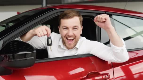 Happy man sits in new car in shop dealership and celebrate purchase of new vehicle. The man with keys shows emotions of happiness while driving in her new car — Vídeos de Stock