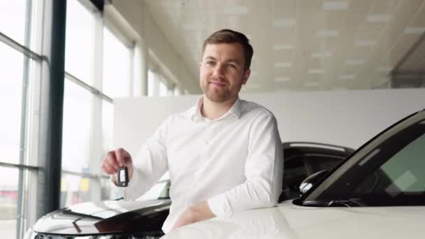Portrait of happy adult successful man posing in auto showroom buying new automobile. Positive male smiling for camera and demonstrating keys while standing near new vehicle in showroom — стоковое видео