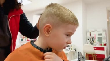 Berber dükkanında saçlarını kuaföre kestiren tatlı, sarışın, gülümseyen bir bebek. Moda uzmanının elleri. Çocuk modası.