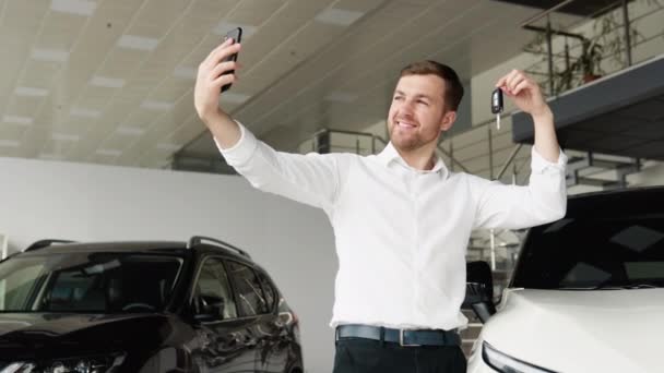 Чоловік щасливо знімає відео біля автомобіля в автосалоні. Щасливий чоловік бере селфі біля своєї нової машини — стокове відео