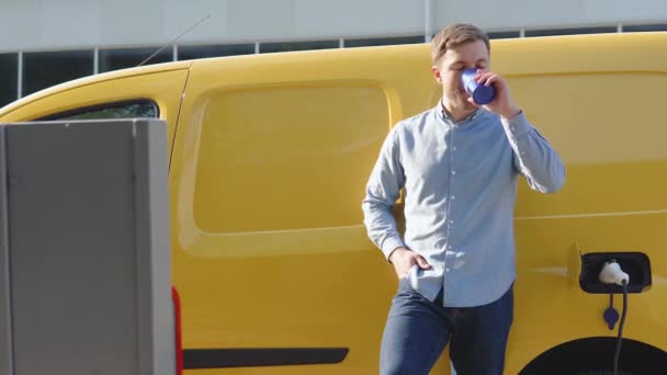 Egy férfi kávét iszik, miközben feltölt egy elektromos autót egy töltőállomáson. Elektromos autós töltés — Stock videók