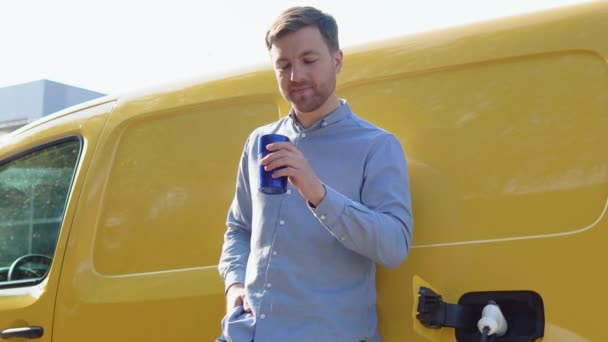Um homem bebe café enquanto carrega um carro elétrico em uma estação de carregamento — Vídeo de Stock