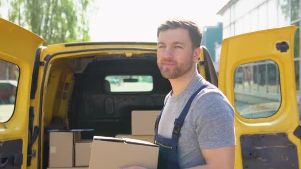 The courier delivers the parcel and hand it to the client. Portrait of a courier holding a parcel, a yellow car in the background — Video