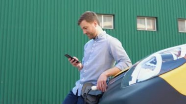 Elektrikli araba istasyonda şarj olurken genç adam cep telefonuyla sohbet ediyor. Yeşil enerji kavramı