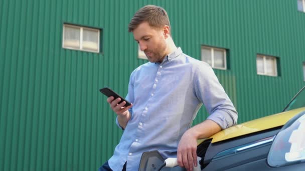 Jongeman chatten op mobiele telefoon terwijl elektrische auto is opladen op outdoor station — Stockvideo