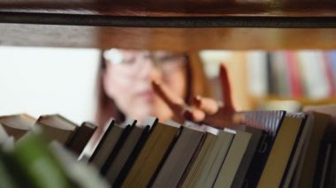 Bir üniversite öğrencisi kütüphanedeki kitaplıklarda bir kitap arıyor. Kütüphanede kitaplıkta kitap seçen bir kız öğrencinin yakın çekimi.