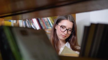 A thoughtful young student reads a book standing in the library. A smiling young girl in glasses reads a book in the university library