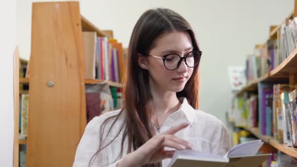 Ένα νεαρό κορίτσι με γυαλιά διαβάζει ένα βιβλίο στη βιβλιοθήκη του πανεπιστημίου. — Αρχείο Βίντεο
