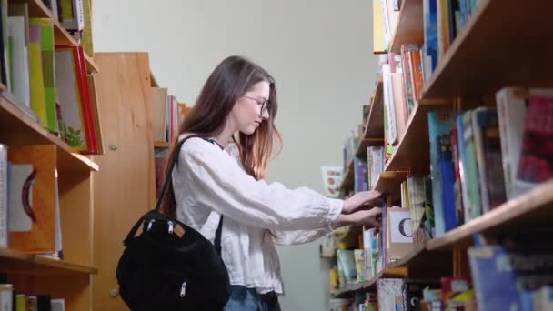 Een universiteitsstudent is op zoek naar een boek op boekenplanken in de bibliotheek — Stockvideo