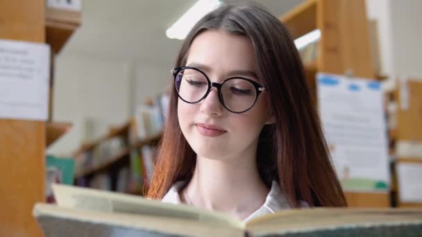 Gözlüklü gülümseyen genç bir kız üniversite kütüphanesinde bir kitap okuyor. Düşünceli genç bir öğrenci kütüphanede ayakta duran bir kitap okuyor. — Stok video