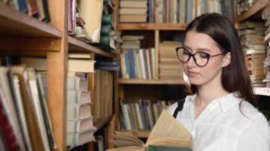 A young attractive student in glasses turns pages in the book, which she took from books on shelves in the library