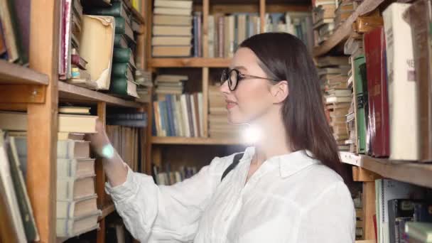 Ein Student sucht in den Bücherregalen der Bibliothek nach einem Buch. Der Student, der den Staub vom alten Buch weht — Stockvideo