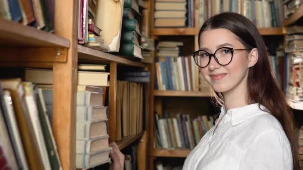 Portrait of a beautiful girl in glasses among the book shelves of the library or archive — Stock video