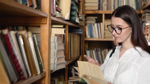 Молода приваблива учениця в окулярах перетворює сторінки в книгу, яку вона взяла з книг на полицях бібліотеки — стокове відео