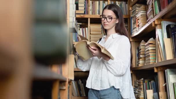 Young attractive student in glasses turns pages in the book, which she took from books on shelves in the library — Vídeos de Stock