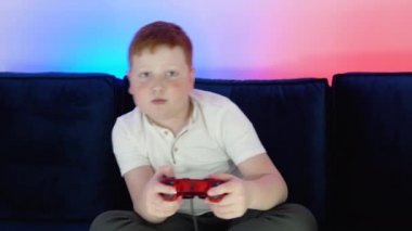 Heyecanlı küçük çocuk oyuncu bir kanepede oturuyor, oyun oynuyor ve konsoldaki video oyunlarında kazanıyor. Sıcak ve neon ışıklı rahat bir oda.