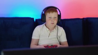 Gamer discussing tactics with teammates while talking into headset. Excited little boy gamer is sitting on a couch, playing and winning in video games on console