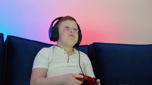 Excited little boy gamer is sitting on a couch, playing and winning in video games on console. Gamer discussing tactics with teammates while talking into headset — Vídeo de stock