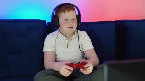一个男孩的游戏玩家坐在监视器前的沙发上，在游戏机上玩着游戏，并在游戏中获胜。有暖气和霓虹灯的舒适房间 — 图库视频影像