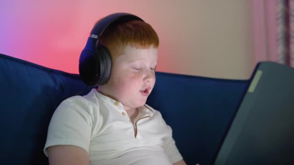 年轻的红头发男生头戴耳机，带着手提电脑视频，与老师进行在线在线在线学习学习。社交距离在家学习语言。家庭教育的概念 — 图库视频影像