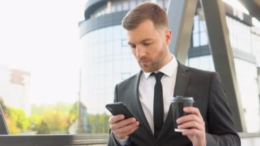 A young confident businessman with a coffee cup using a mobile phone near a business center