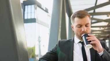 Kendine güvenen genç işadamı kahve içiyor ve iş merkezinin yakınındaki kameraya bakıyor.